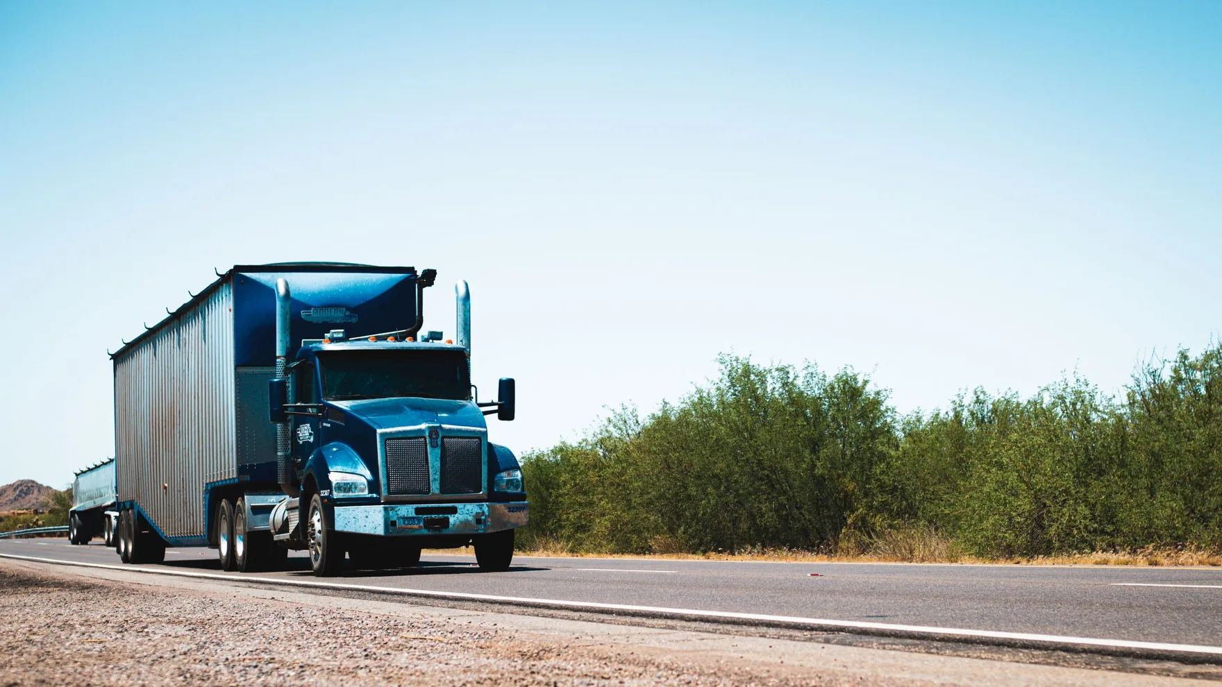 A blue semi truck hauling a trailer travels along an open highway under a clear sky.