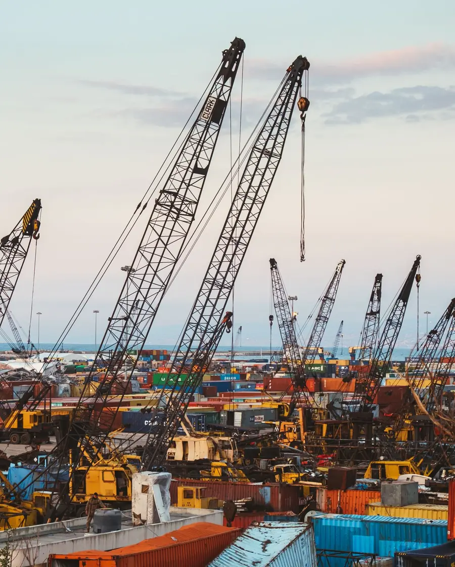 Portside cranes and stacked cargo containers at an industrial harbor with heavy machinery in the foreground.