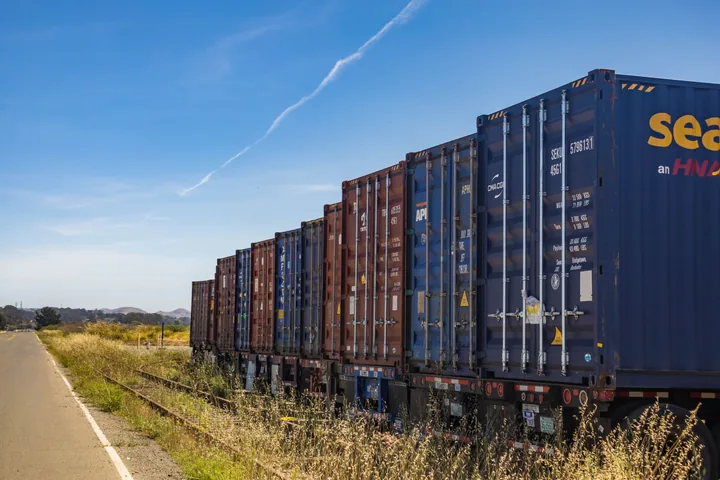 A freight train hauling stacked cargo shipping containers on railway tracks under a blue sky.