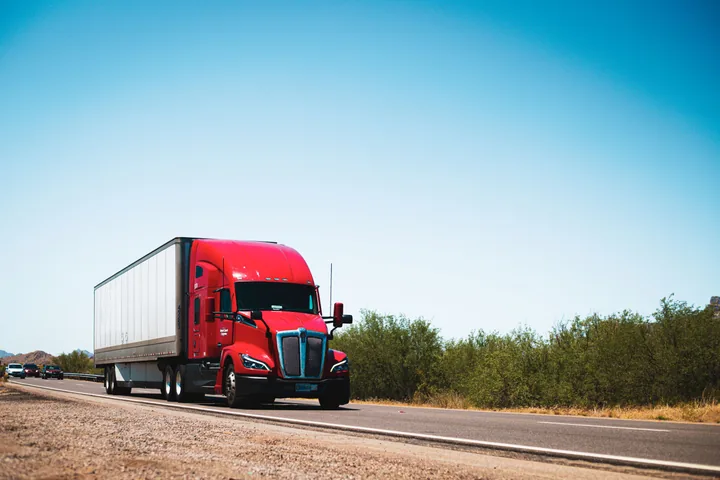 Aerial view of a container semi truck driving along a road beside a green forest.