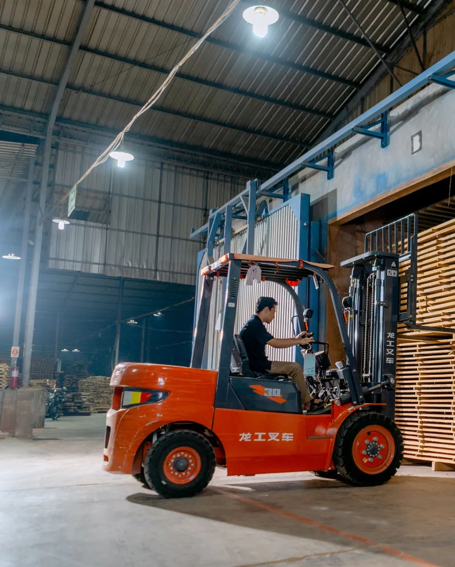 A worker drives a forklift lifting wooden pallets among tall storage racks inside a warehouse.